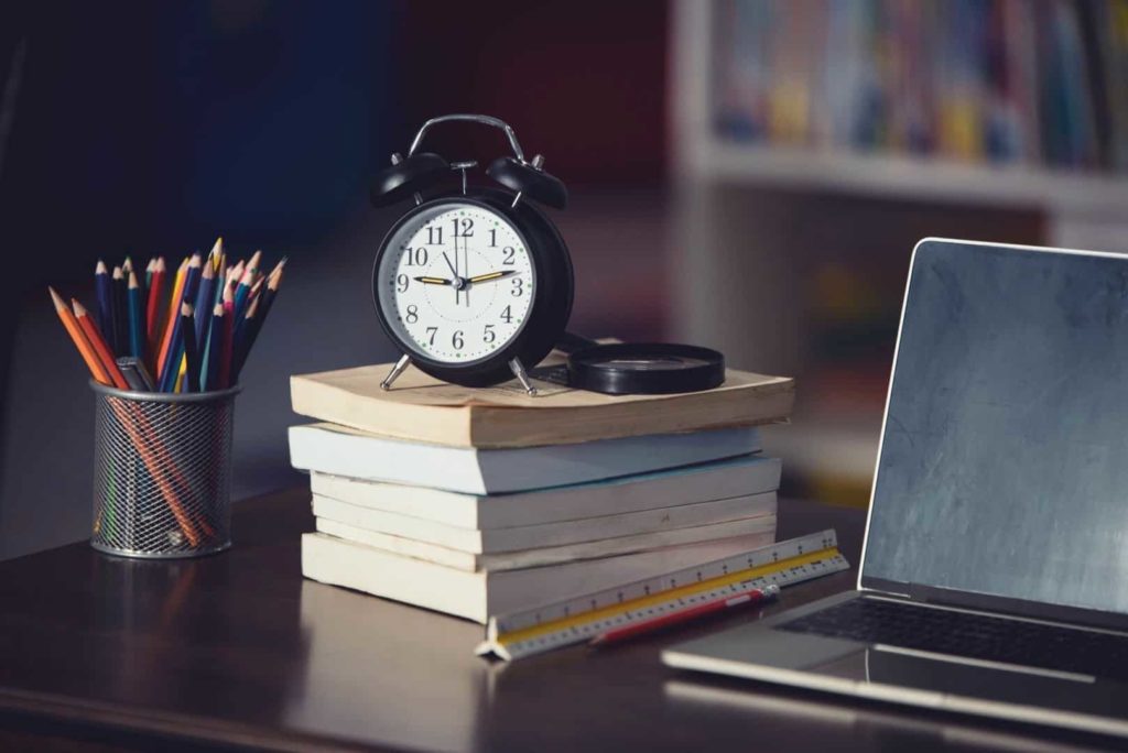 A clock is on the pile of books near the laptop and pencils